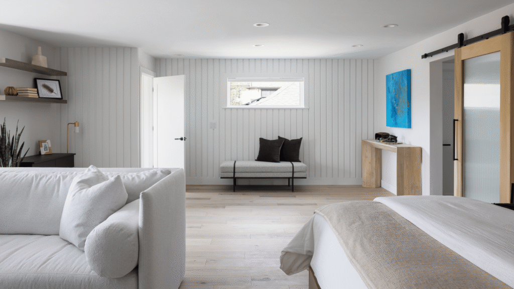 Bright primary bedroom suite with white oak floors, white furnishings, vertical wall paneling, and a sliding barn door.